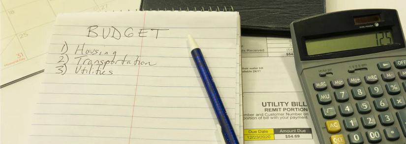 A photo of a calculator, a utility bill, a calendar, pen, and notepad. On the notepad is Budget and a list of items.