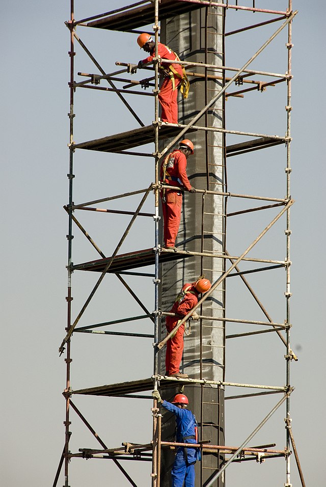 Four workers in helmets and uniforms work at different levels on a tall scaffolding structure.