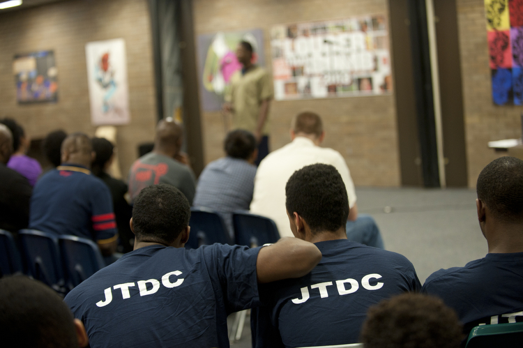 Individuals in a room listening to someone, some wearing prison uniforms. 